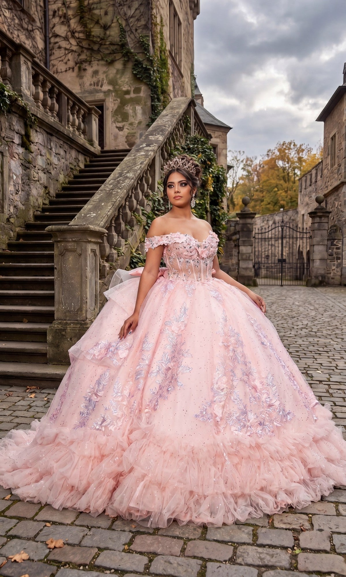 A woman wearing the Quinceanera Dress CHF3141 By Chicas in blush, featuring 3D floral accents and a corset-style bodice, paired with a tiara, stands on a cobblestone path near stone stairs and historic buildings.